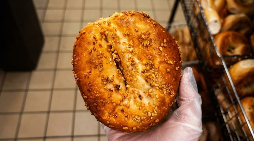 Gloved hand holding a freshly baked, golden-brown bagel topped with crunchy garlic bits.