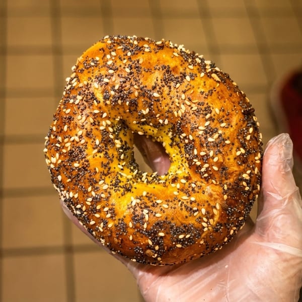 A gloved hand holds up a freshly baked everything bagel, covered in poppy and sesame seeds.