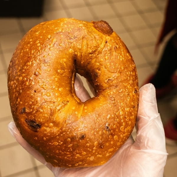 A gloved hand holds a golden-brown bagel with a slightly bumpy texture, against a tiled floor background.