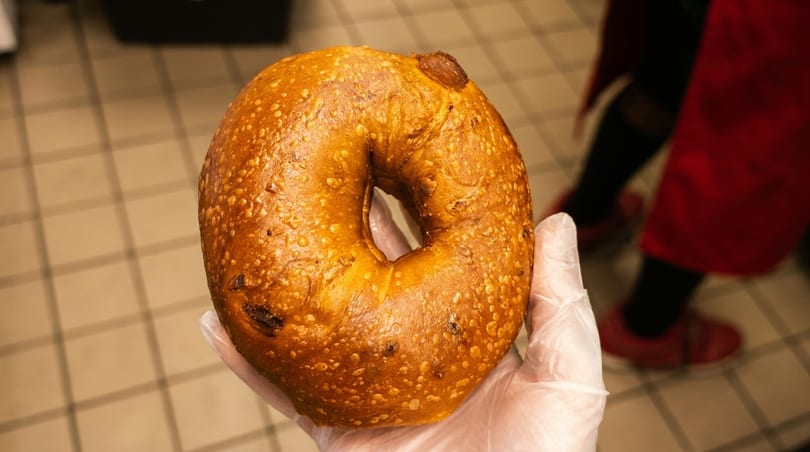 A gloved hand holds a golden-brown bagel with a slightly bumpy texture, against a tiled floor background.