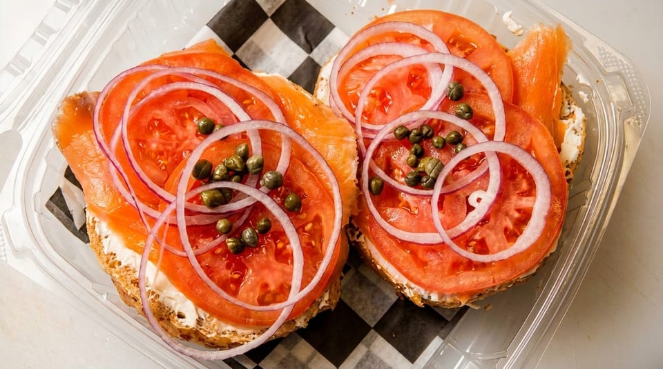 Two bagels with cream cheese, smoked salmon, tomato, red onion, and capers in a plastic container.