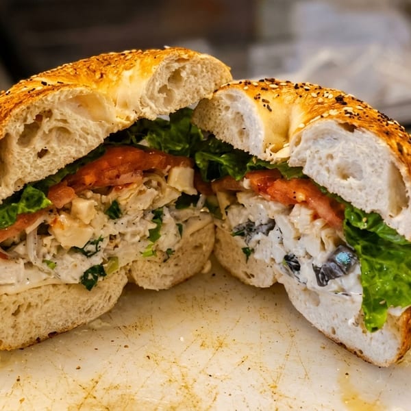 A bagel sandwich, cut in half, filled with chicken salad, lettuce, and tomatoes.